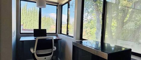 A room with a desk, chair, television, and large windows offering a view of trees and mountains.