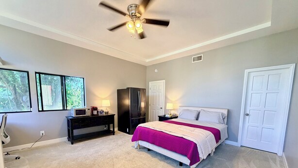 A bedroom with a bed, a ceiling fan, a refrigerator, a nightstand, a lamp, and a door. - Peaceful Foothill Getaway Tennis & 40+ Fruit Trees (Upland)