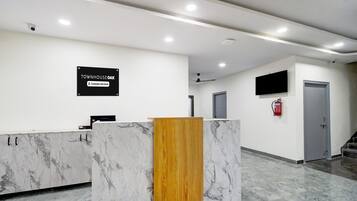 Una recepción con mostrador de mármol, panel de madera, cartel que indica 'Company Serviced', extintor, televisor y una escalera.