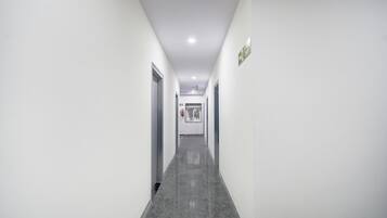 Um corredor comprido e estreito com paredes brancas e chão cinzento, que leva a uma porta no final.