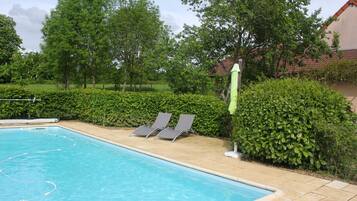 A swimming pool with clear blue water, surrounded by a tiled deck. There are two lounge chairs and a hedge along the poolside.
