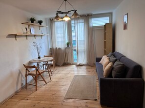 Ein gemütliches Wohnzimmer mit einem dunklen Sofa, Holzboden und einem runden Tisch mit Stühlen.
