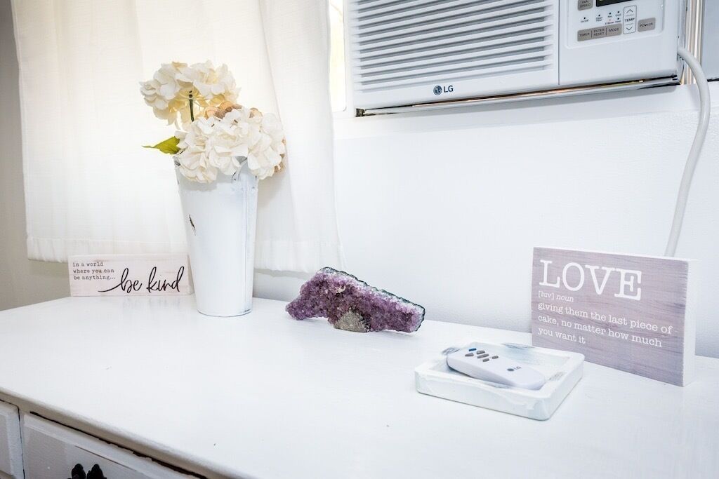 A white bedside table with a vase of flowers, a crystal, a remote control, and a sign with the word 'LOVE'.