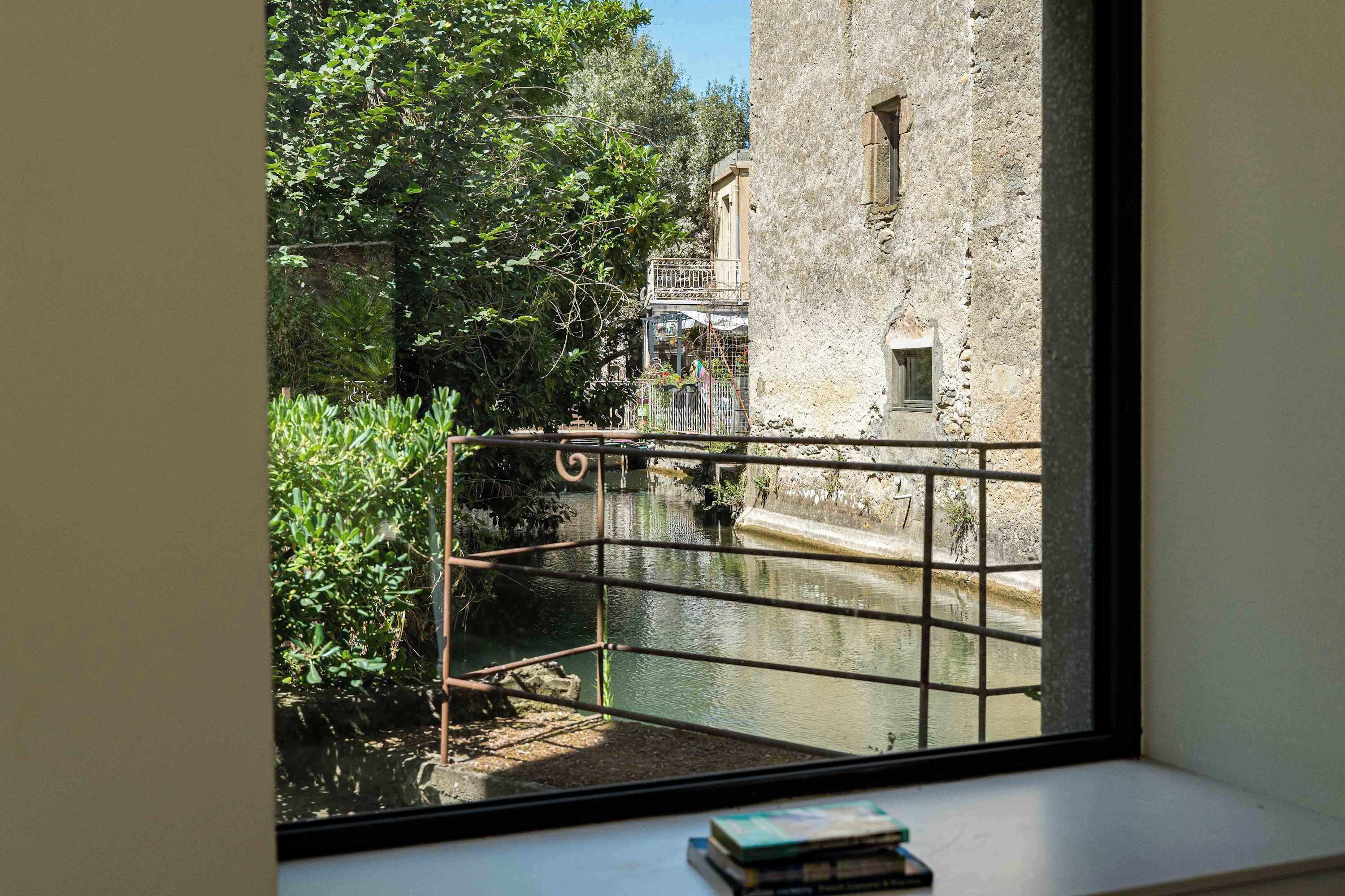 Vista a través de una ventana que muestra un canal, follaje y un muro de piedra.