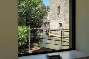 Blick durch ein Fenster auf einen Gebäudebereich mit Wasserkanal, Grünflächen und einer Steinmauer.