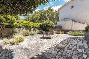 Un patio con piso de piedra, una mesa con sillas y un edificio blanco al fondo.