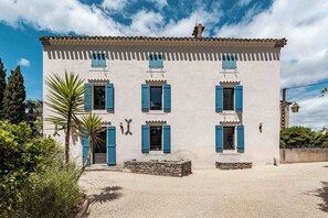 A two-story building with a symmetrical facade, multiple windows with blue shutters, and a stone wall in front. - Mill near Carcassonne, with a pool and river (Bouilhonnac)