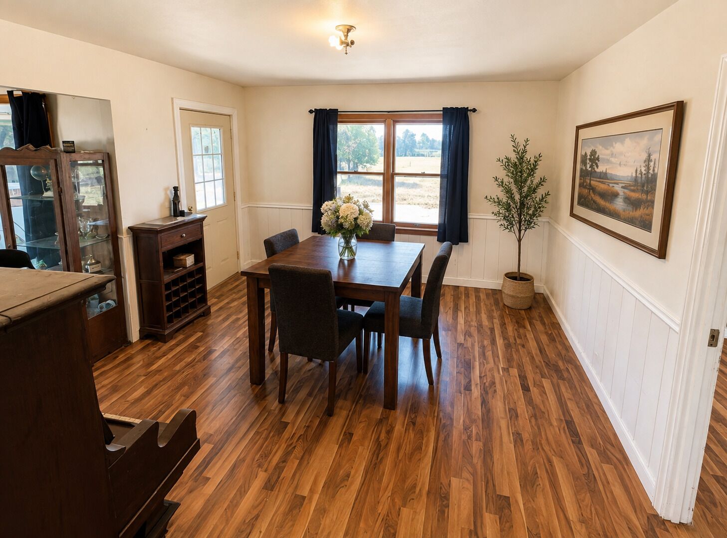 A dining area with a wooden table, chairs, a vase of flowers, and a window with curtains.
