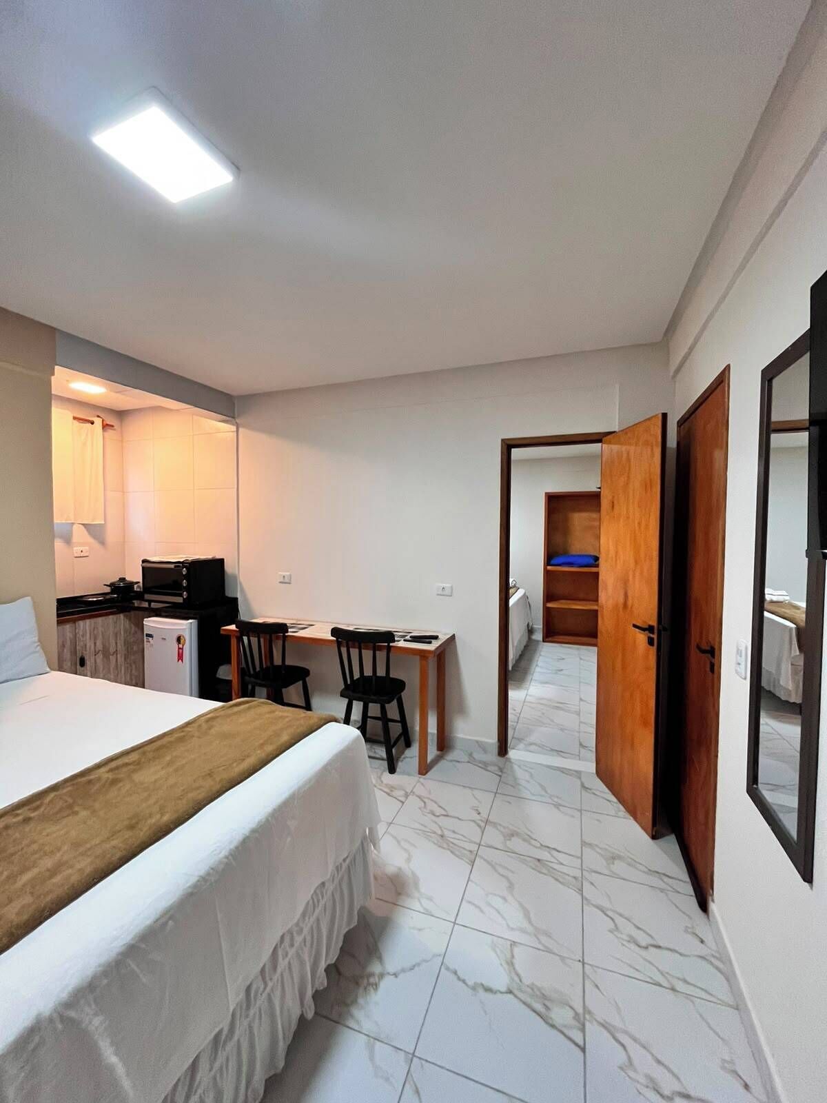 A hotel room with a bed, a desk, a chair, a small kitchenette, and a bathroom.