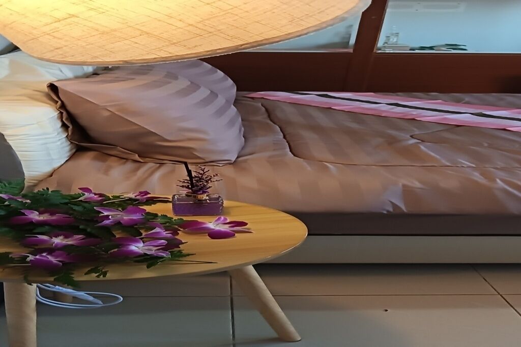A bed with a beige cover, a wooden nightstand with a lamp, and a small vase with flowers.