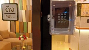 Zona de recepción de un hotel con puerta de cristal, rótula de emergencia, mostrador de recepción y un muro decorativo.