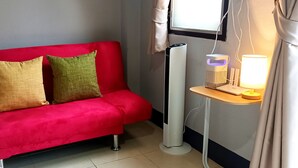 A red sofa with two pillows, a standing heater, and a bedside table with a lamp.