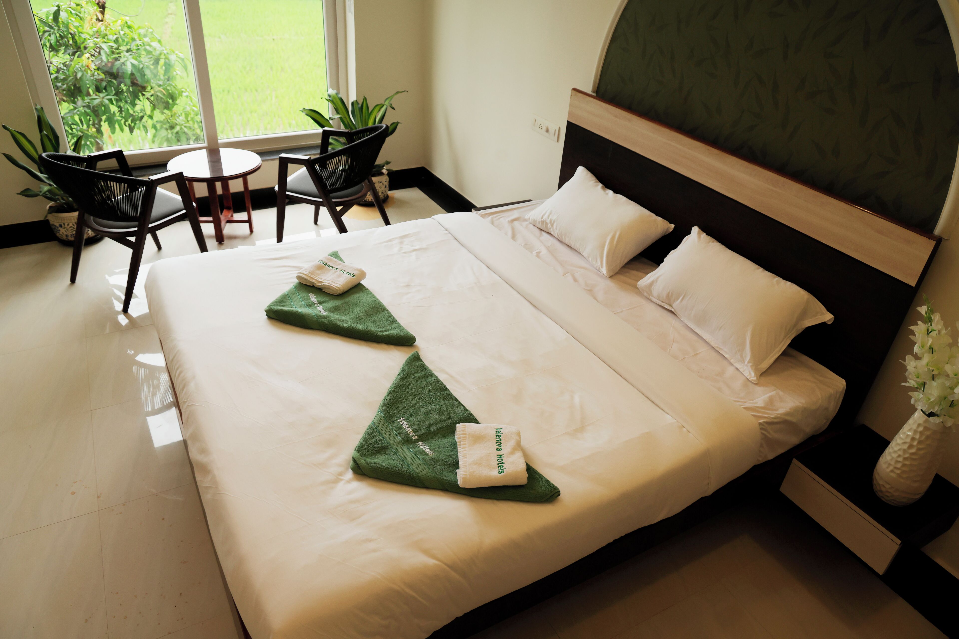 Quarto de hotel com uma cama, duas almofadas, uma toalha verde e um vaso com flores.