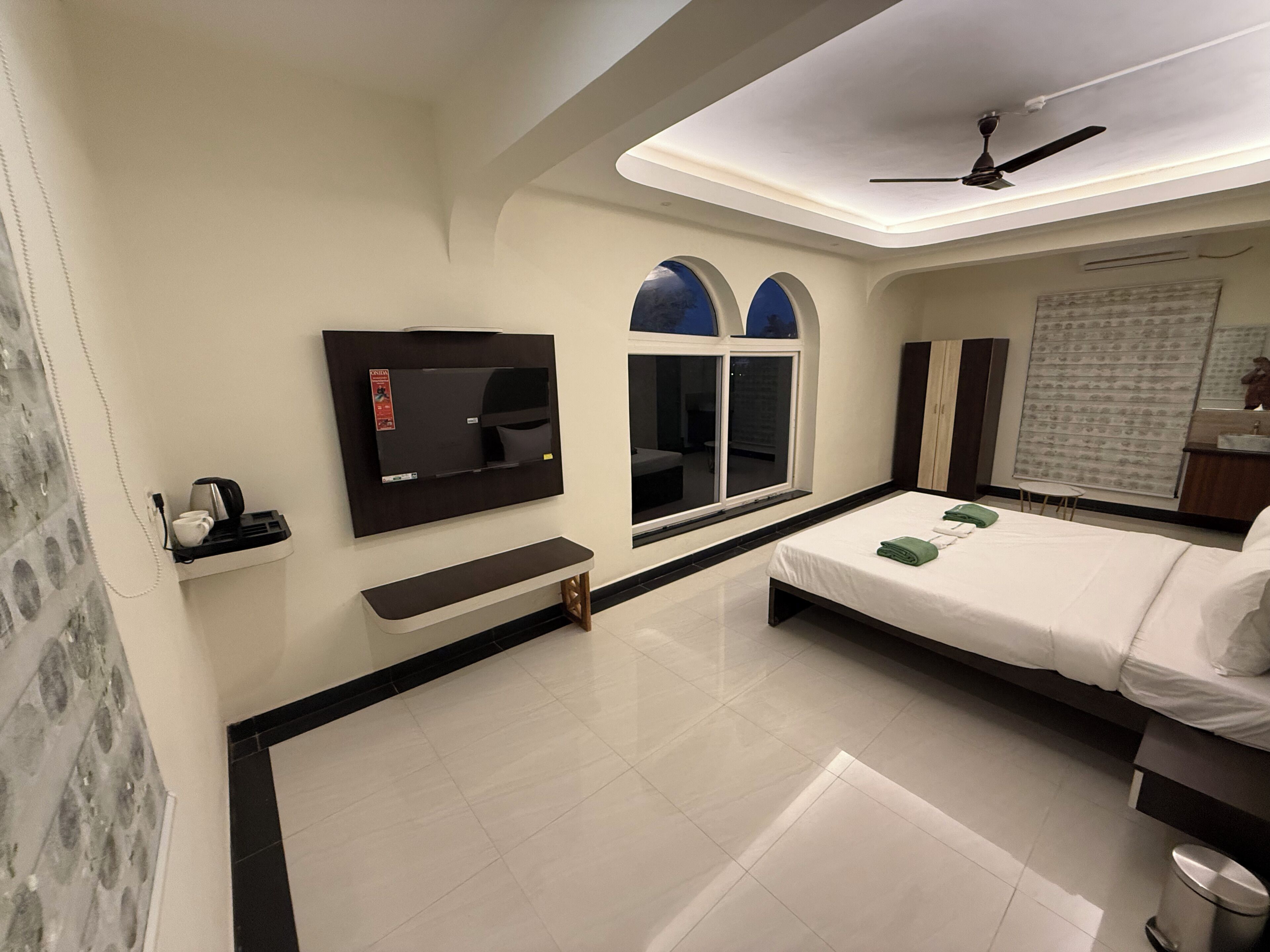 Una habitación de hotel con cama, televisor montado en la pared, ventana y ventilador de techo.