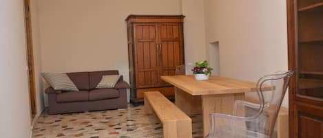 A small room with a wooden dining table, a bench, a clear plastic chair, a brown sofa with patterned cushions, and a wooden cabinet.
