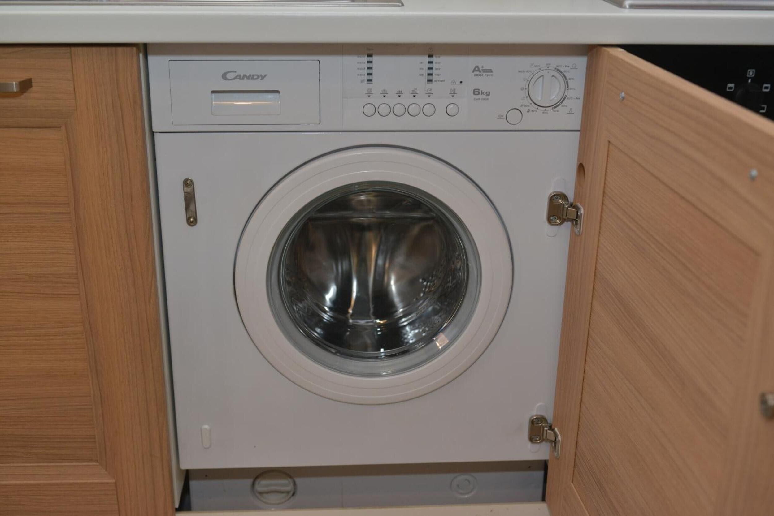 A compact, front-loading washing machine with a control panel, located in a wooden cabinet.