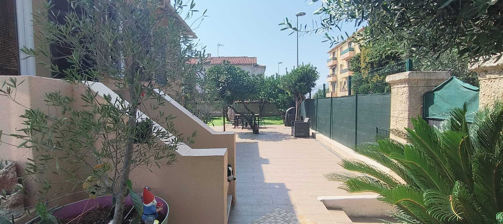 Un chemin pavé mène à un bâtiment entouré d’une clôture verte et bordé d’arbres.