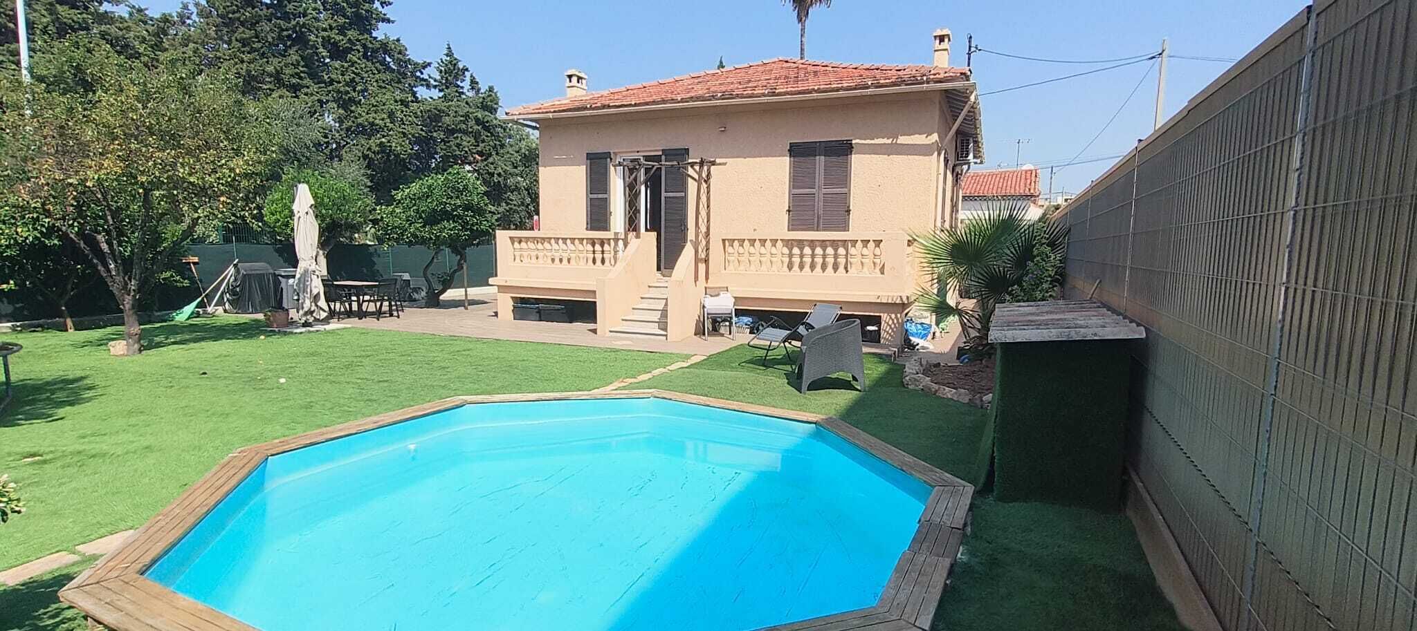 A swimming pool with a wooden deck, a house with a red-tiled roof, and a fenced yard with grass and trees.