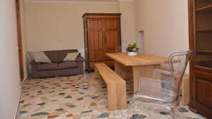 Une petite pièce comprenant une table à manger en bois, un banc, une chaise en plastique transparent, un canapé marron avec des coussins à motifs et un meuble en bois.