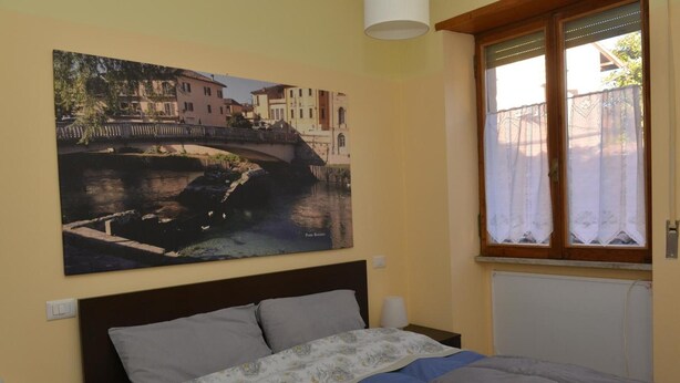 Une chambre à coucher avec un lit, un tableau représentant un pont, une fenêtre avec des rideaux et un radiateur.