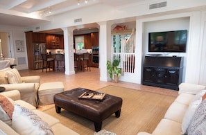 A spacious living room with a large TV, a leather ottoman, and a kitchen area in the background. - BREATHTAKING : on water front with panoramic views + pool + dock (Dunmore Town)