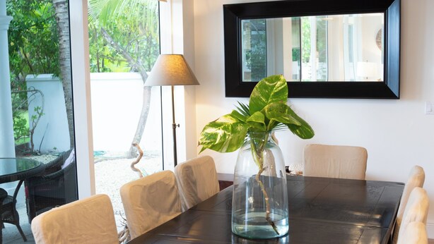 A dining area with a dark wooden table, white cushioned chairs, a glass vase with a plant, a ceiling fan, and a large window with a view of greenery. - BREATHTAKING : on water front with panoramic views + pool + dock (Dunmore Town)