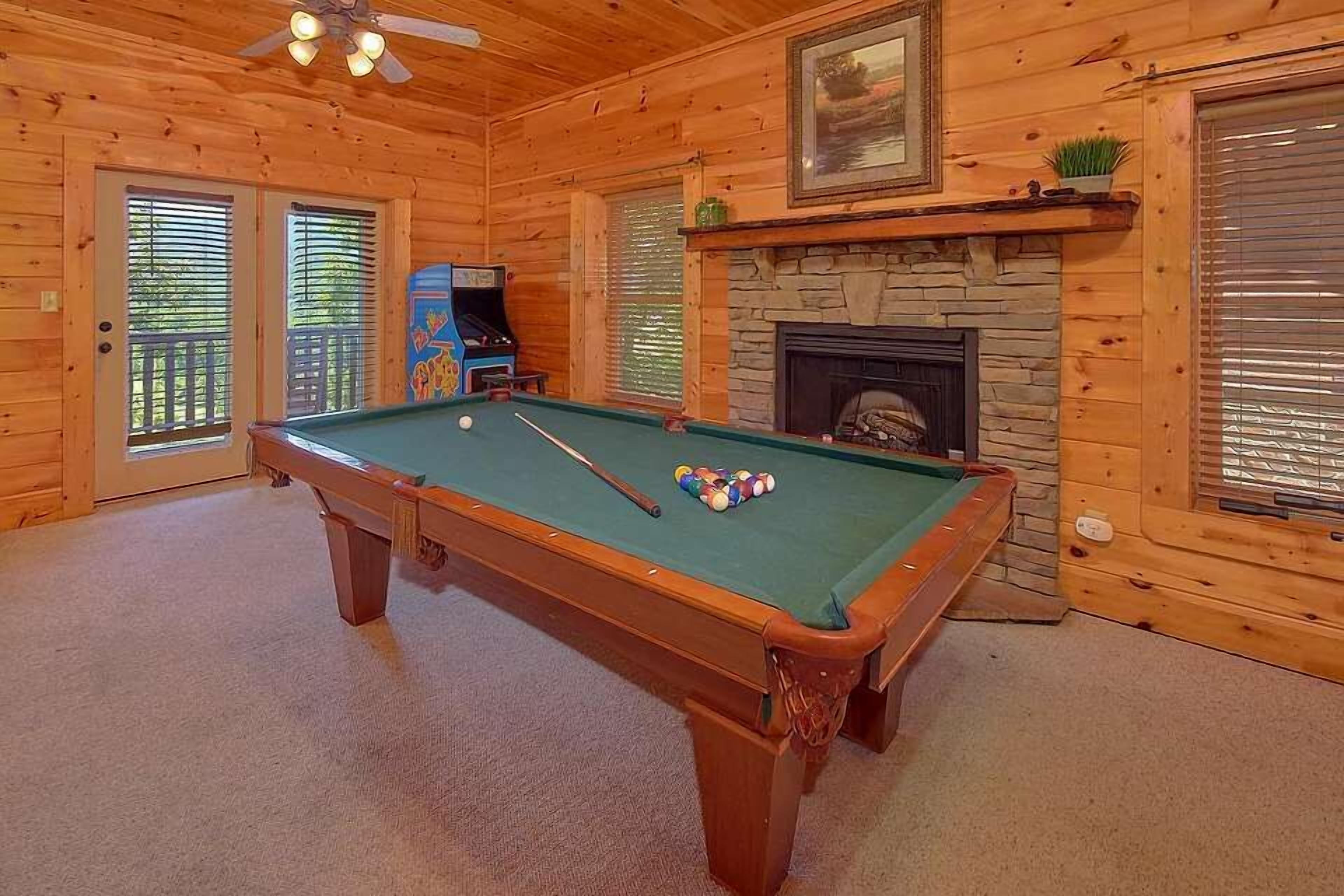 A pool table in a wooden room with a fireplace and arcade games.