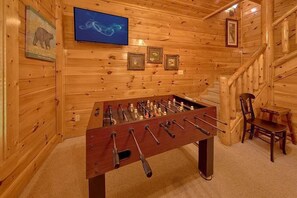 Habitación con piso de madera, mesa de fútbol de mesa, televisor montado en la pared y cuadros enmarcados.