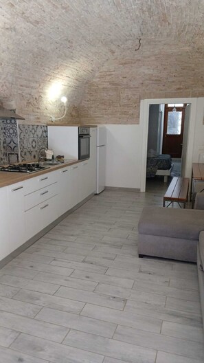 A kitchen with white cabinets, a stove, and a refrigerator. There is a wooden bench and a sofa in the living area.