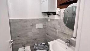 A compact bathroom with a toilet, bidet, and sink, featuring grey tiles and wooden accents.