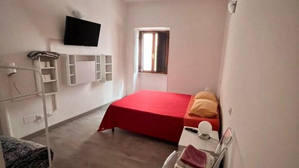 A compact bedroom with a bed, a TV mounted on the wall, a window, and a small table with a towel.