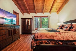 Une chambre avec un plafond en bois, un grand lit avec une literie à motifs, une commode, un téléviseur et une fenêtre donnant sur des arbres.
