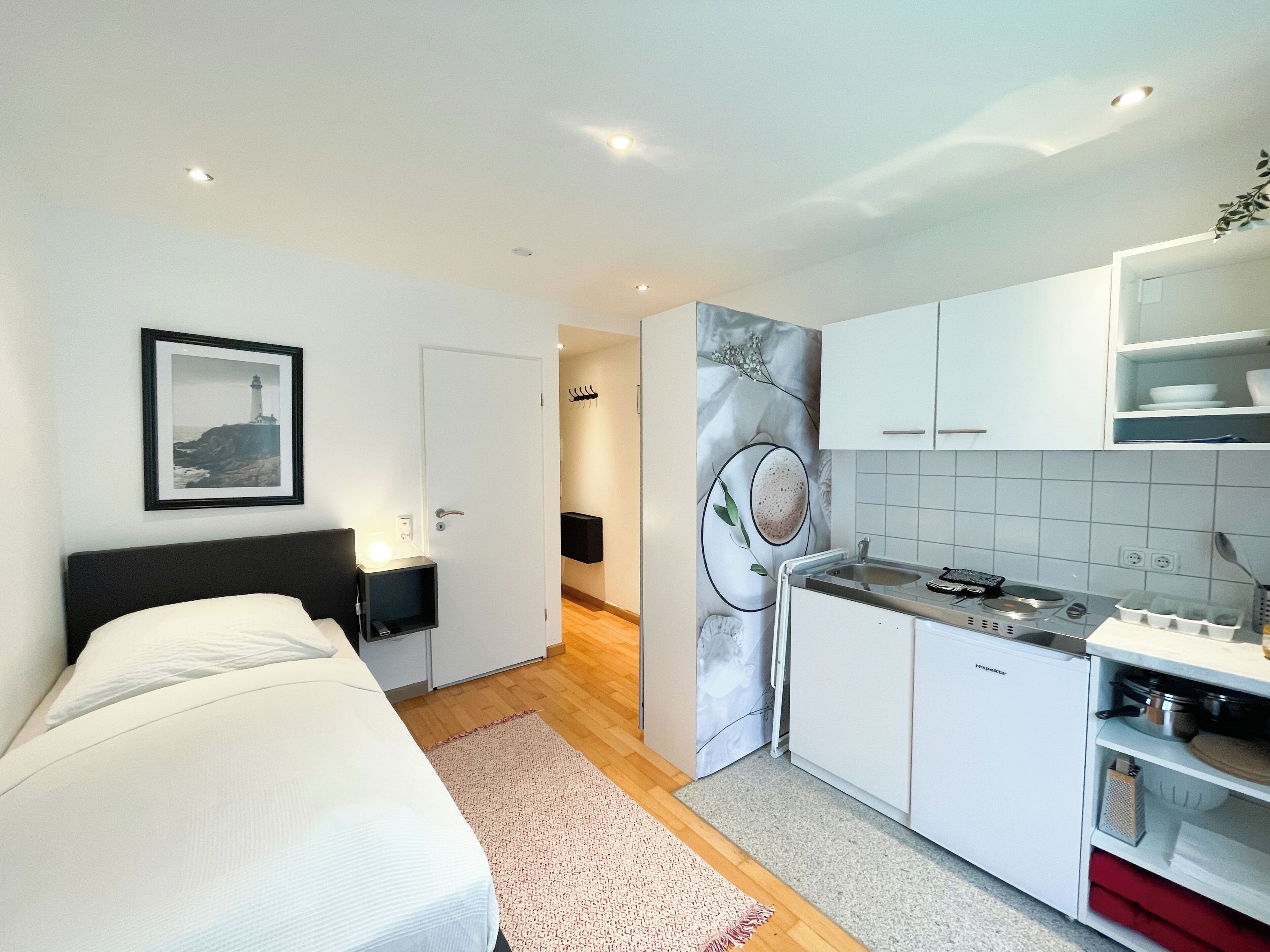 Ein modernes Schlafzimmer mit Bett, Nachttisch und Beistelltisch. Durch eine offene Tür ist ein Badezimmer mit Dusche und Toilette zu sehen. Der Raum hat weiße Wände, einen Holzboden und einen Küchenbereich mit weißen Schränken und einer Arbeitsfläche.