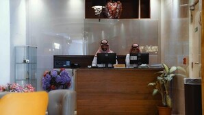 Un comptoir d’accueil avec deux employés, une enseigne indiquant « réception », une plante en pot et un espace d’attente avec une chaise.