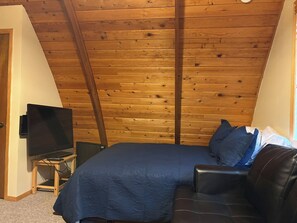 A bedroom with a sloped ceiling, a bed with a blue bedspread, a black leather sofa, and a television on a wooden stand. - Cozy & Secluded Forest Staycation-BBQ (Brighton)