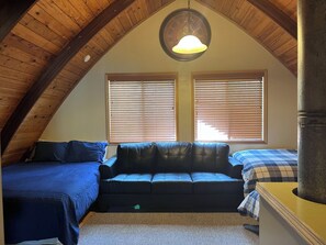 A bedroom with a sloped ceiling, a black leather sofa, two beds with blue bedding, and a round wall clock. - Cozy & Secluded Forest Staycation-BBQ (Brighton)