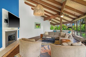 A living room with a fireplace, comfortable seating, a ceiling fan, and large windows offering a view of greenery. - Mauna Kai 7B by Gather | Princeville 3BR + Loft Condo w/ Shared Pool (Princeville)
