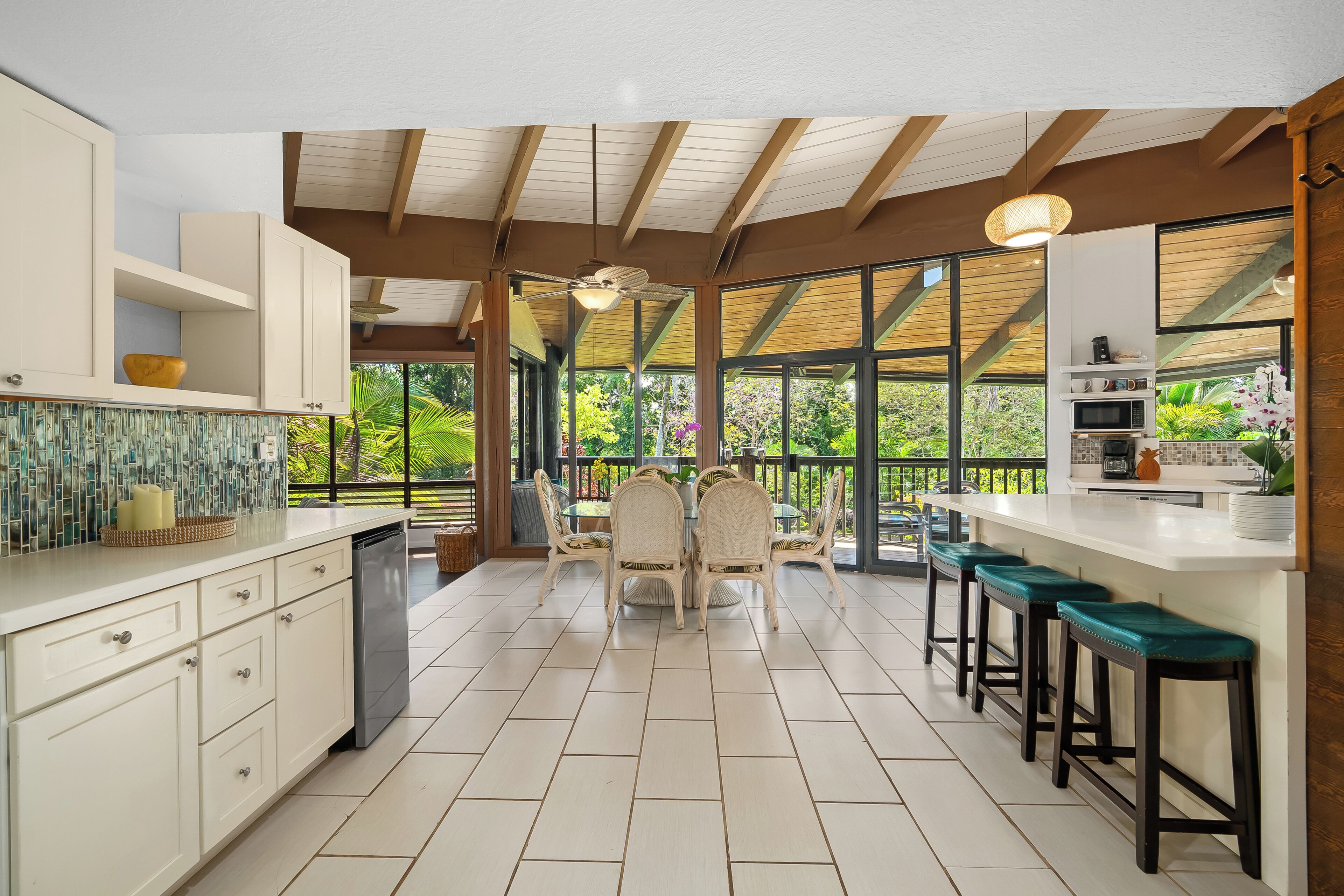A modern kitchen with white cabinetry, a dining area with rattan chairs, and large windows offering a view of greenery.