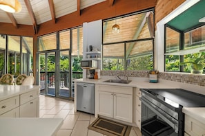 A modern kitchen with a dishwasher, microwave, and stove, large windows, and a view of greenery. - Mauna Kai 7B by Gather | Princeville 3BR + Loft Condo w/ Shared Pool (Princeville)