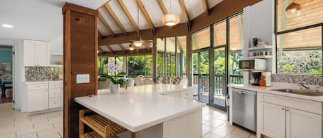 A modern kitchen with a large island, wooden beams, and a view of greenery outside.