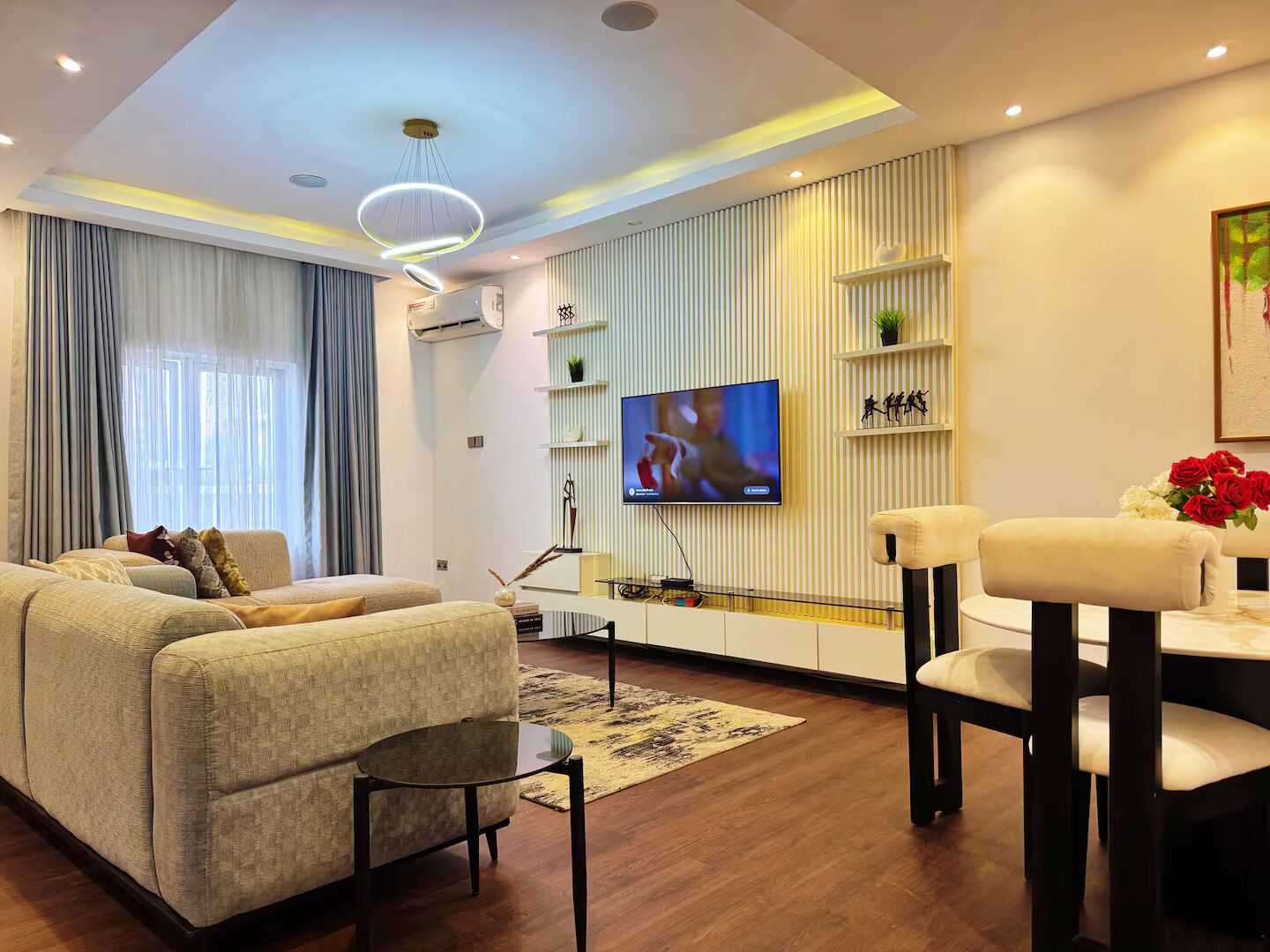 Een moderne woonkamer met een bank, een salontafel, een aan de muur bevestigde televisie en een eethoek met stoelen.