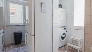 A compact laundry room with a washing machine, a small table, and a trash bin. - 2BR Beachside Living by LovelyStay (Costa da Caparica)