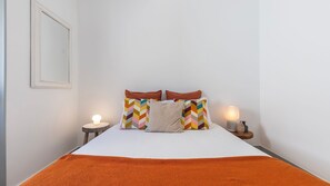 Uma cama de solteiro com colchão branco, duas almofadas com estampas geométricas, um cobertor marrom e duas luminárias de cabeceira sobre mesas de cabeceira de madeira.