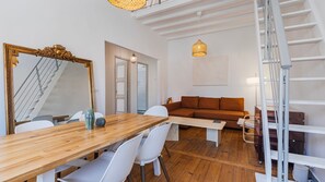 A modern living room with a wooden dining table, white chairs, a brown sofa, a large mirror, and a staircase. - 2BR Beachside Living by LovelyStay (Costa da Caparica)