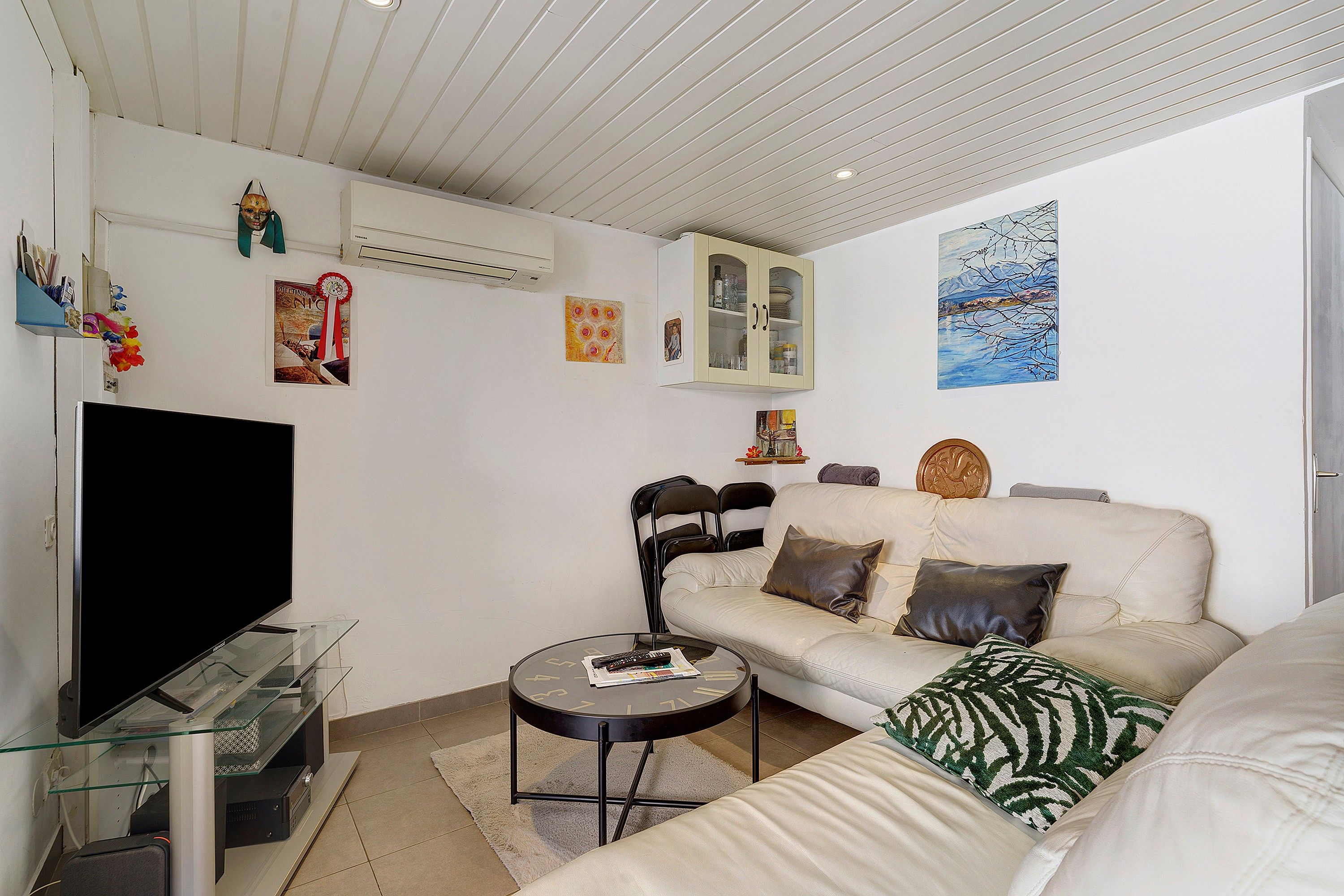 Un salon avec un téléviseur à écran plat, une table basse en verre, un canapé beige et un tableau accroché au mur.
