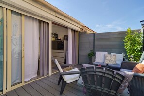 Un espacio de estar techado al aire libre con sillas y una mesa, una puerta corrediza de vidrio que da a una cocina y una valla de madera.