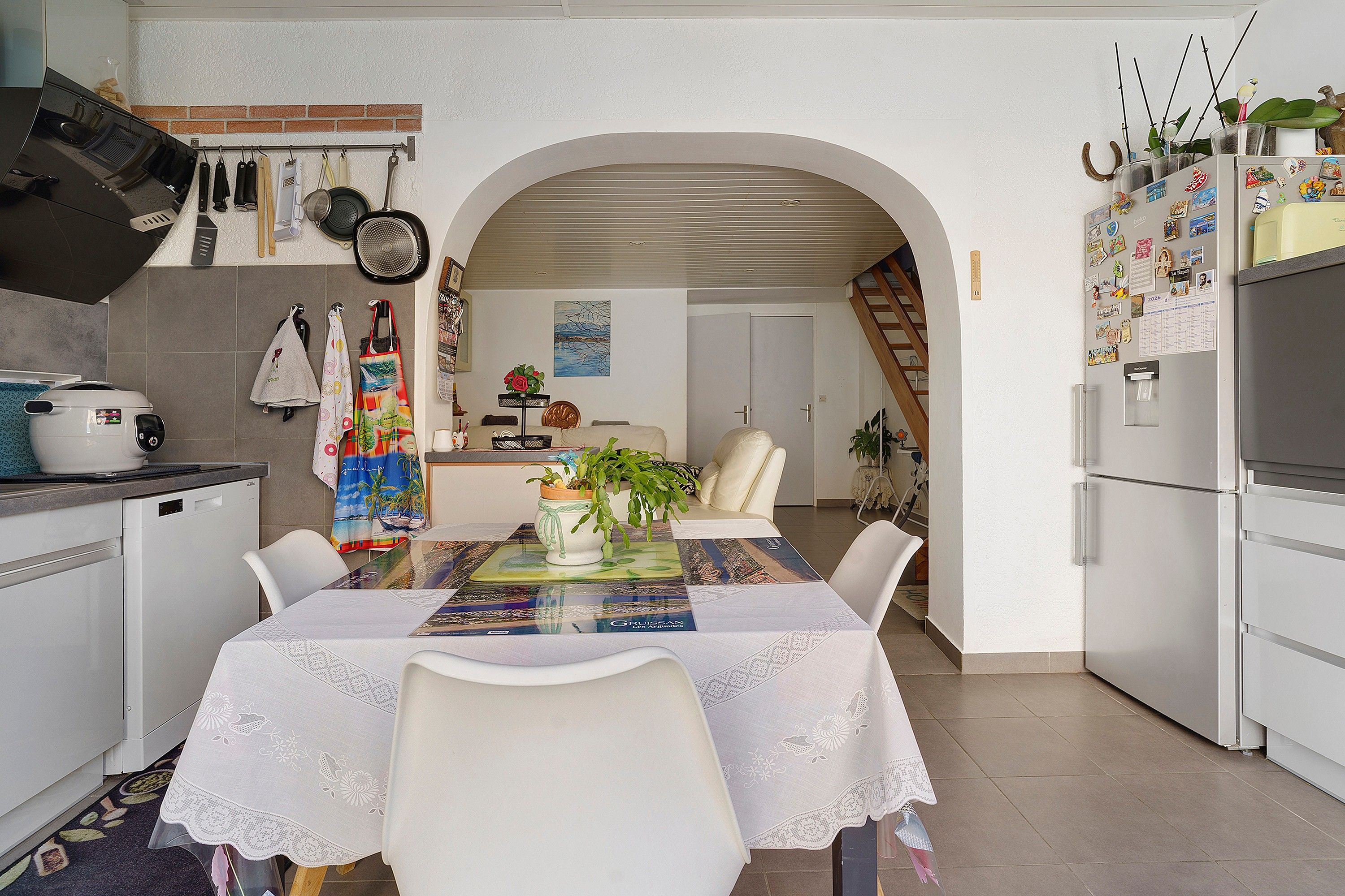 Une cuisine avec des appareils électroménagers blancs, une table à manger agrémentée d’un chemin de table coloré, et une vue sur l’espace repas.