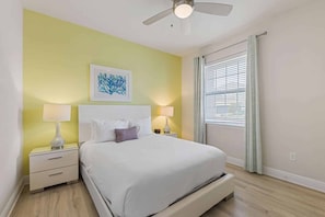 A hotel room with a bed, two bedside tables, a ceiling fan, a window with curtains, and a wall-mounted artwork. - Harmony Palms Villa - Eight Bedroom House, Sleeps 17 (Lake Buena Vista)