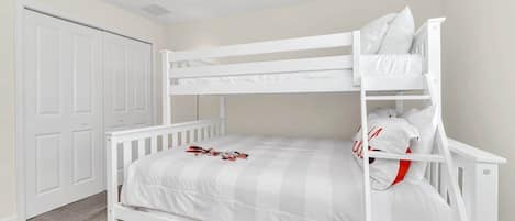 Una cama litera con estructura blanca y ropa de cama gris, una puerta blanca y piso alfombrado.