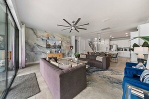Una sala de estar moderna con sofá, mesa de centro y televisor. Se observa una cocina al fondo con armarios blancos y una zona de comedor.